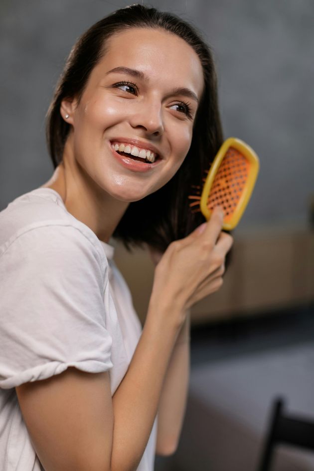 Una persona sonriente con el pelo hasta los hombros usa un cepillo de pelo con mango amarillo en una habitación con una pared gris.