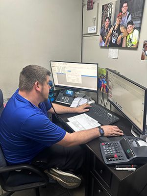 A person in a blue shirt works at a dual-monitor desk in an office with a photograph displayed on the wall. | Military Brake & Alignment Services Inc.
