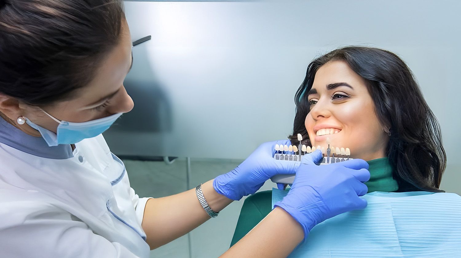 A Dentist Wearing A Mask And Blue Gloves Holds A Tooth Shade Guide Up To A Patient's Smile