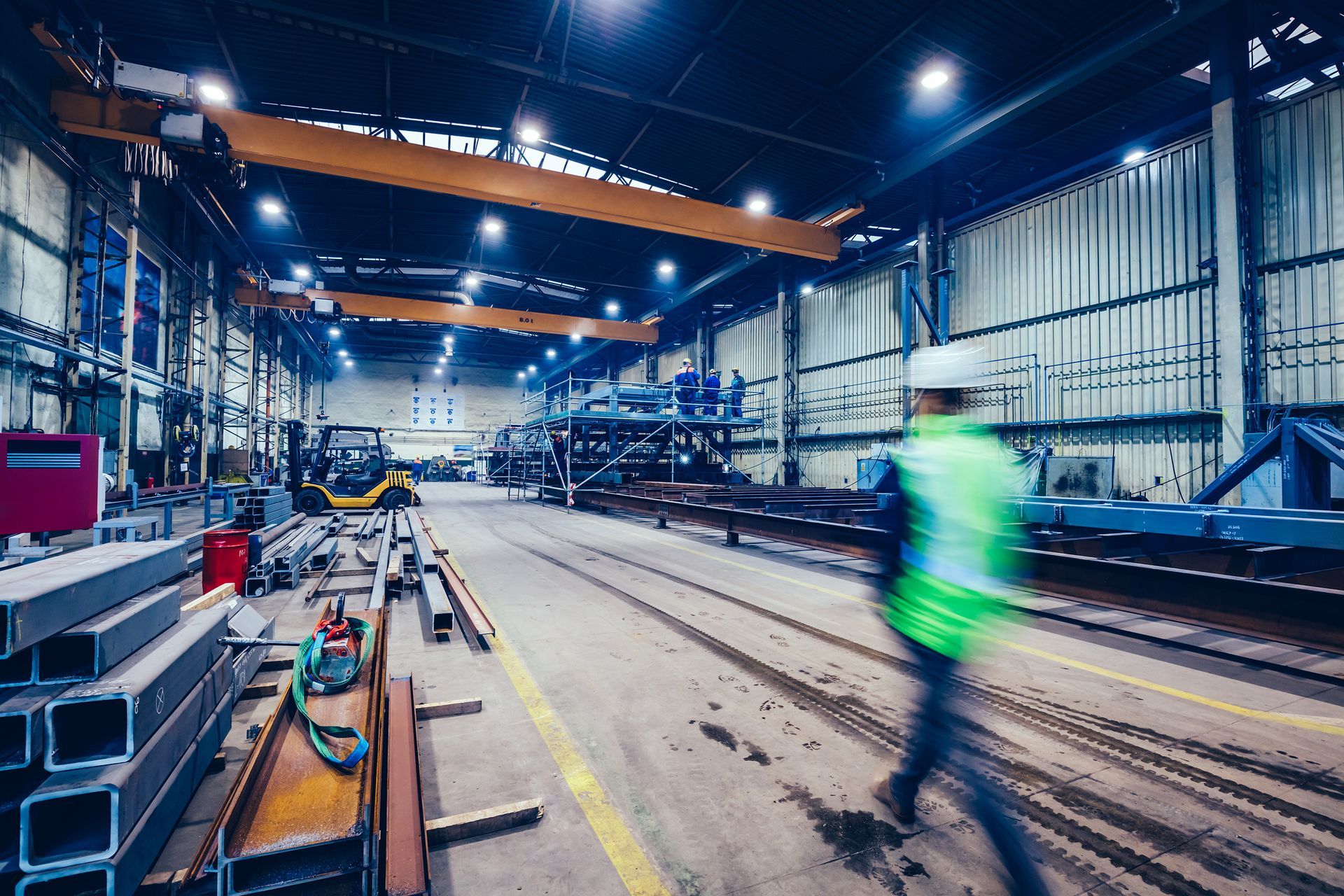 Inside a factory, worker in vest walks past machinery and metal beams under bright lights.
