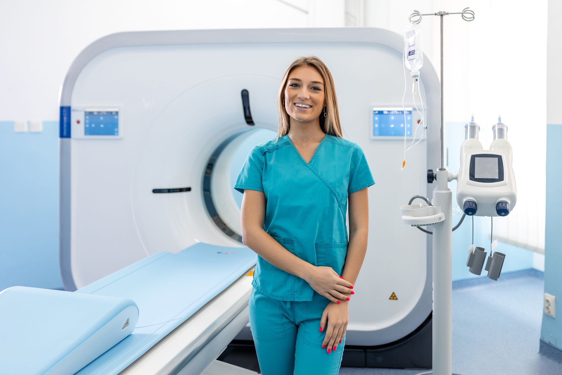 Smiling medical worker in teal scrubs stands by a CT scanner in a hospital room.
