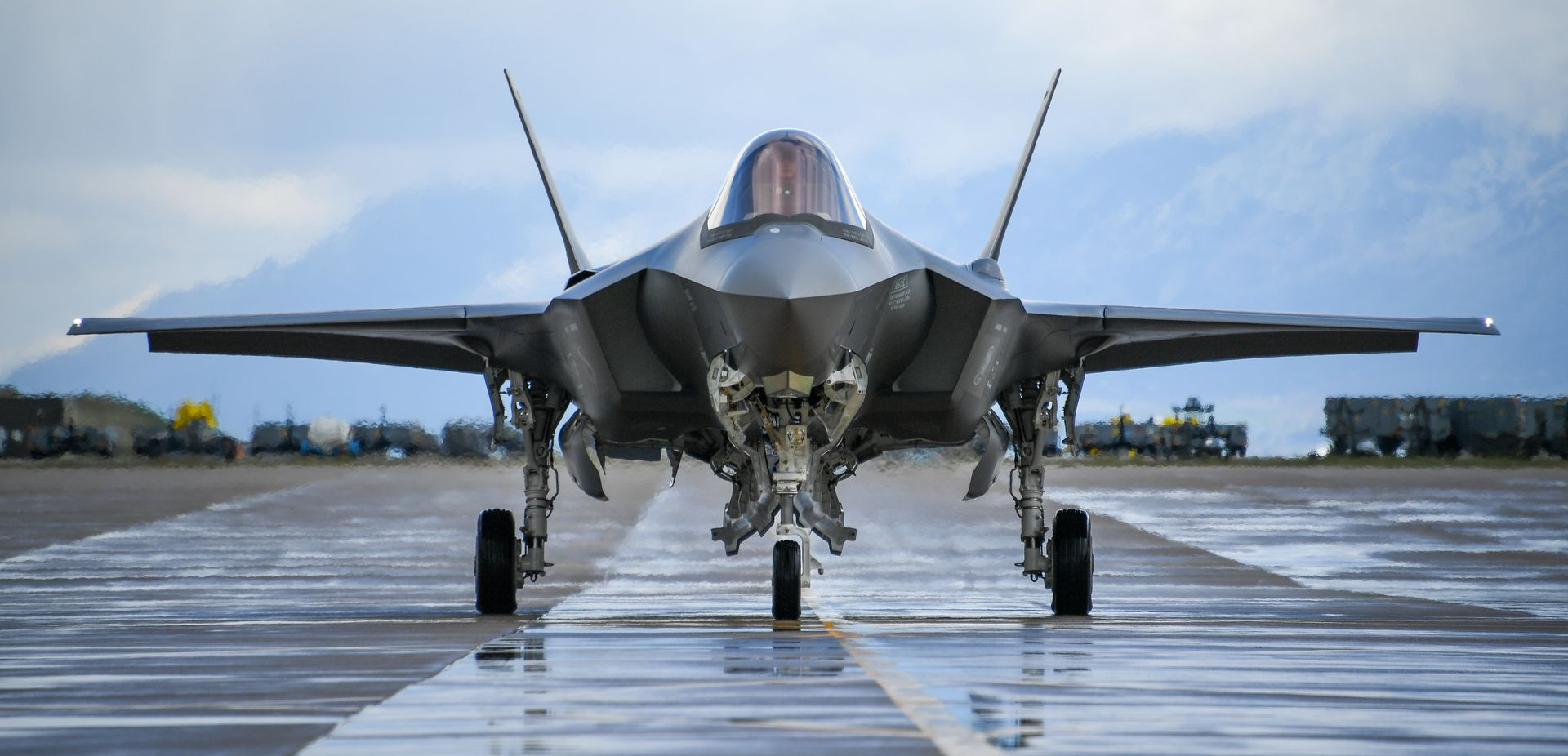 An F-35 fighter jet on a wet runway, with mountains in the background under an overcast sky.
