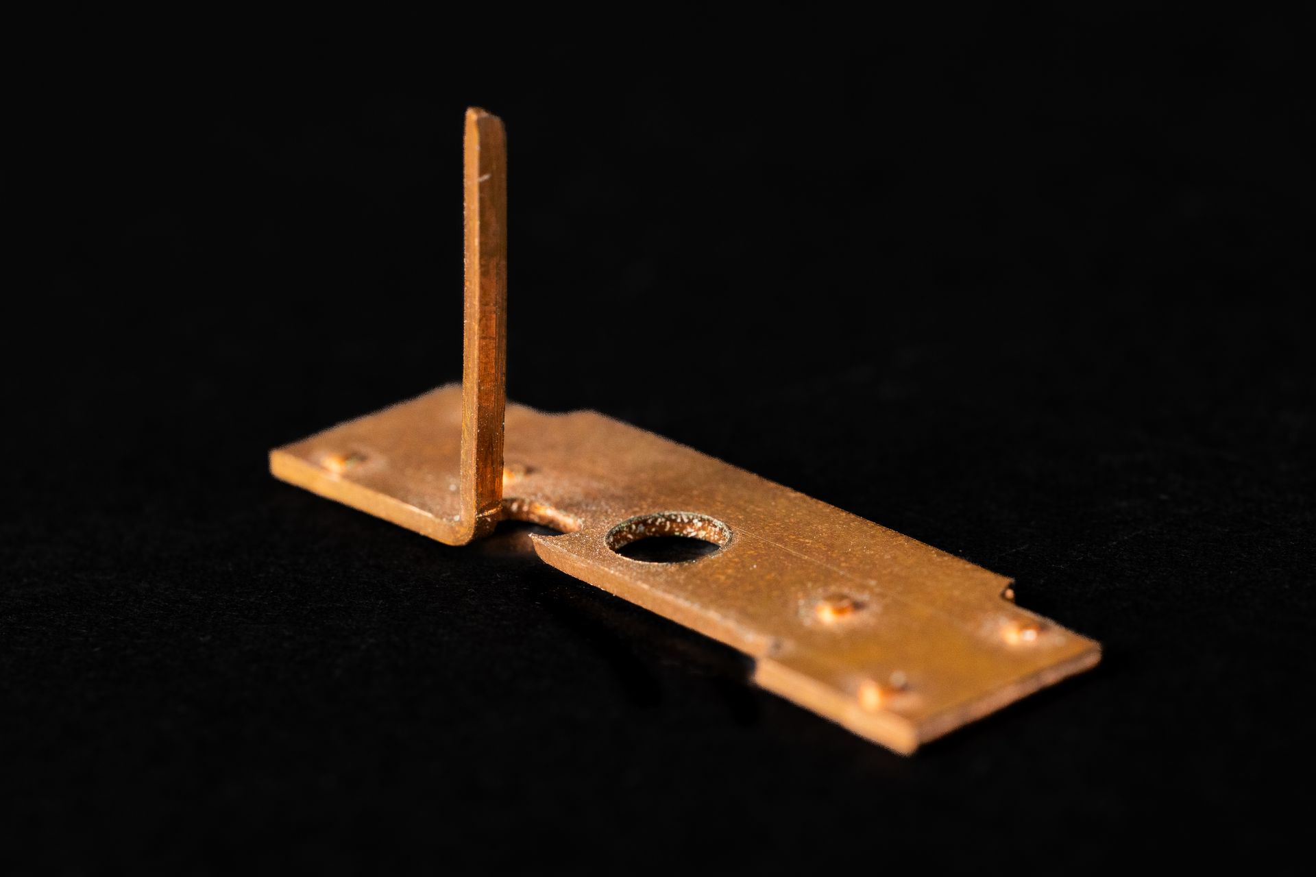 Copper-colored metal bracket with a protruding pin, several holes, and a bent section; set against a black background.