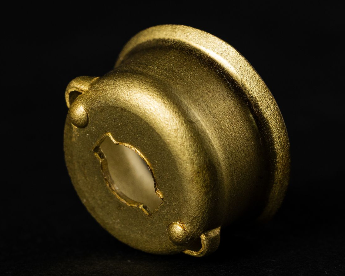 Golden-colored metal vessel with a curved rim, three small handles, and an oval opening, set against a black background.