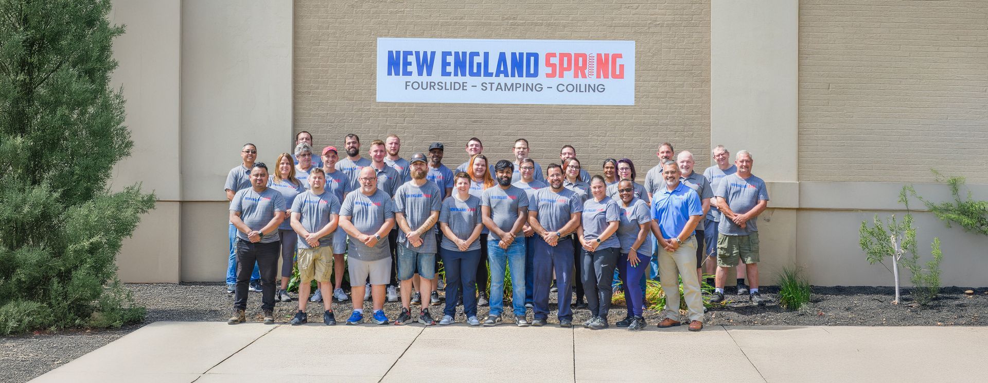 Group photo of New England Spring employees in front of their building. They are wearing gray shirts and khaki or dark pants.