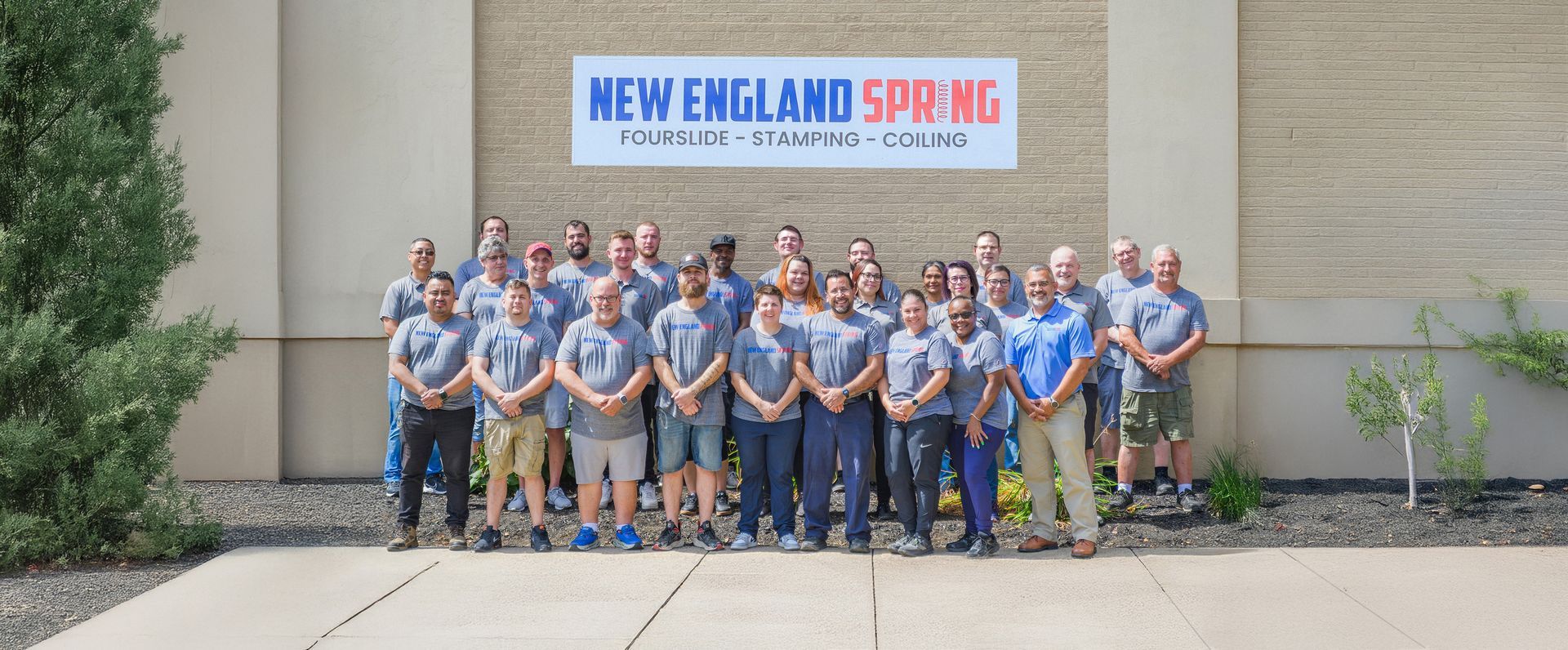 Group photo of New England Spring employees in front of their building. They are wearing gray shirts and khaki or dark pants.