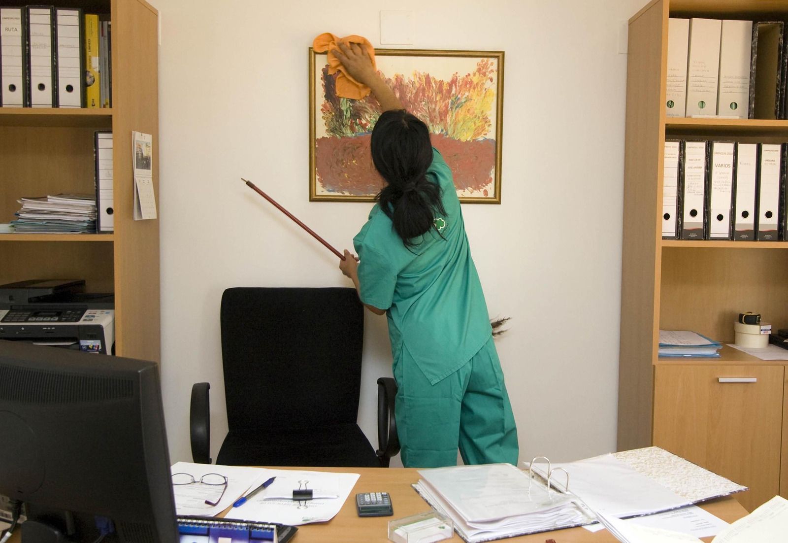 A woman in a green scrub suit is cleaning a picture on the wall