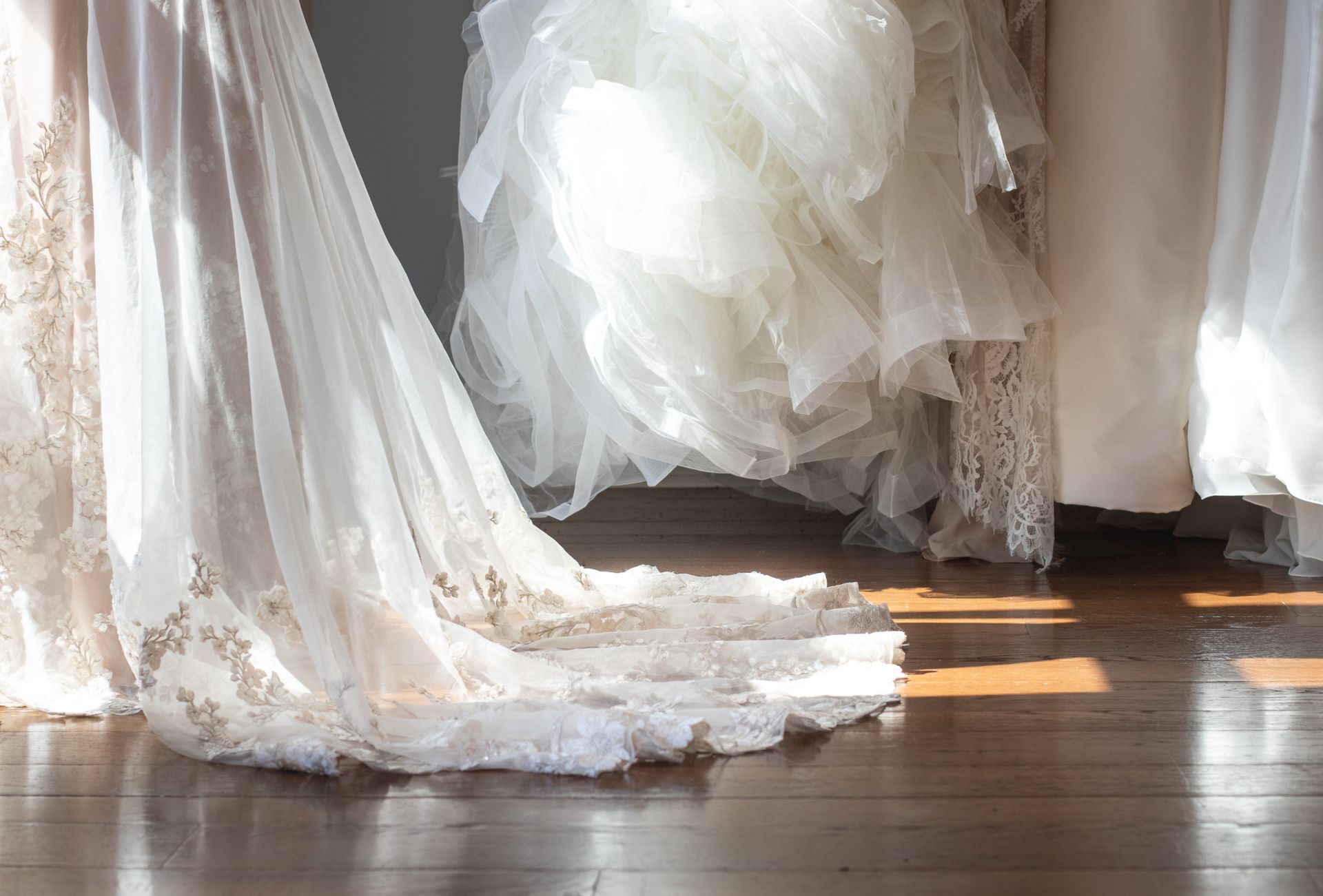 White bridal dresses hang in a bright room, their delicate lace trains draped over a polished wooden floor.