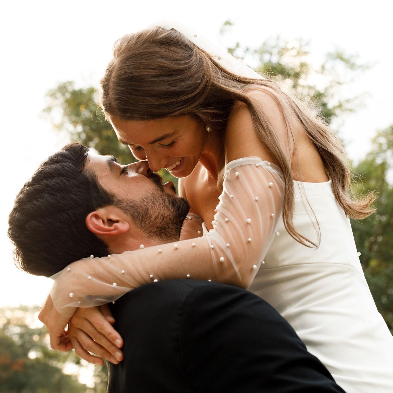 A person in a white gown with pearl-embellished sleeves embraces a person in a black suit, noses touching affectionately.