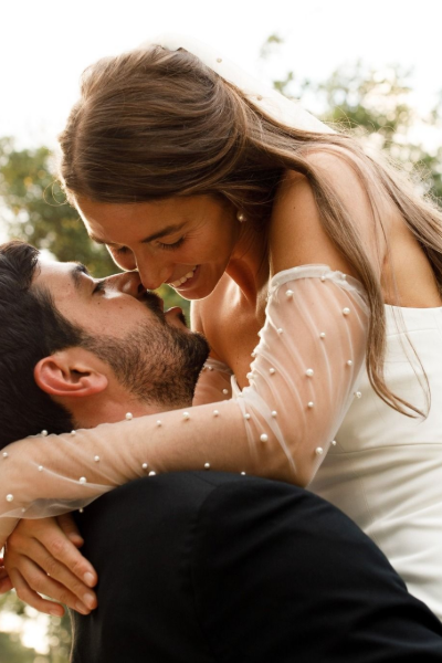 A person in a white gown with pearl-embellished sleeves embraces a person in a black suit, noses touching affectionately.