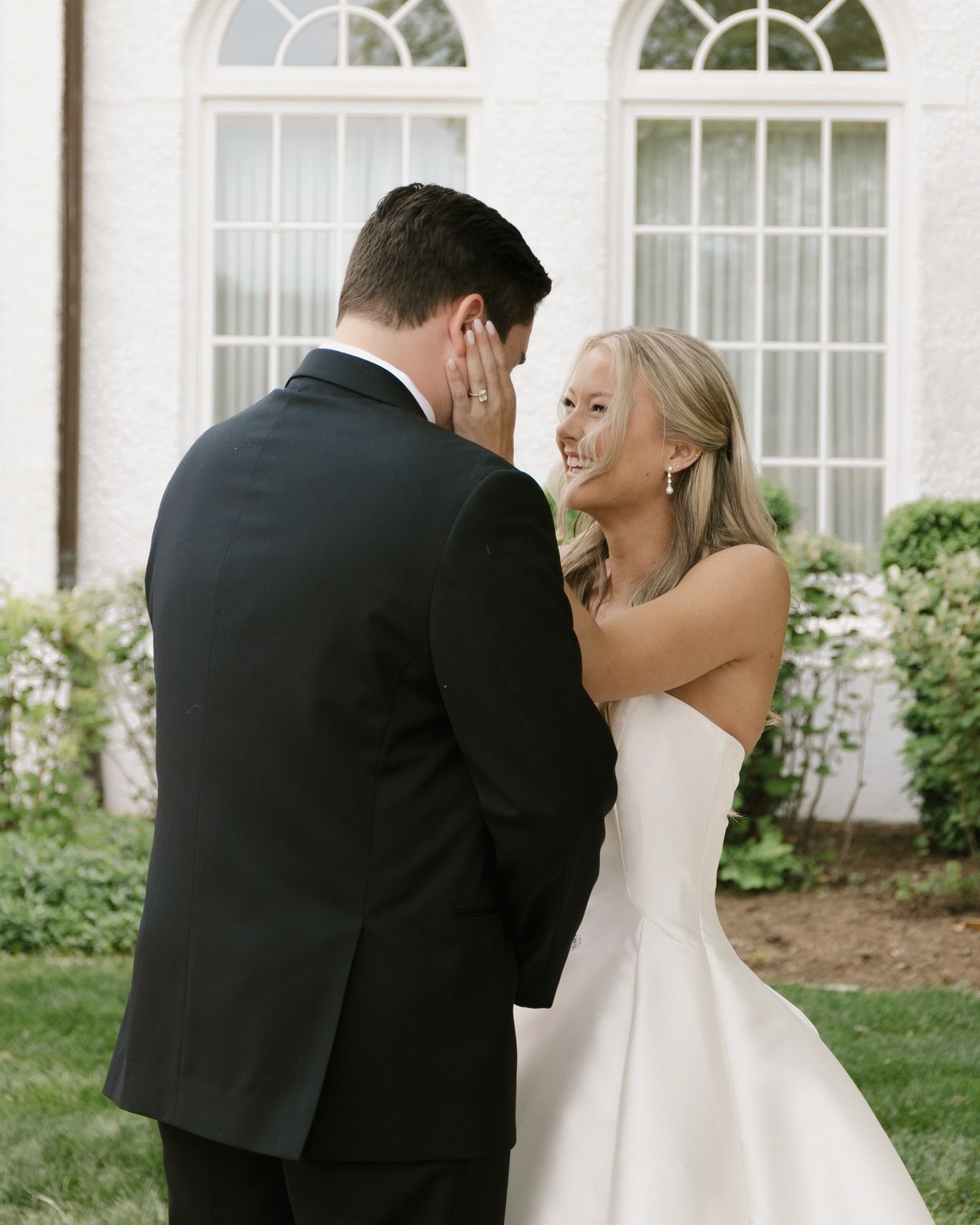 A person in a wedding dress touches the face of a person in a suit while smiling in front of a white building.