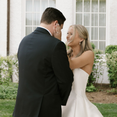 A person in a wedding dress touches the face of a person in a suit while smiling in front of a white building.