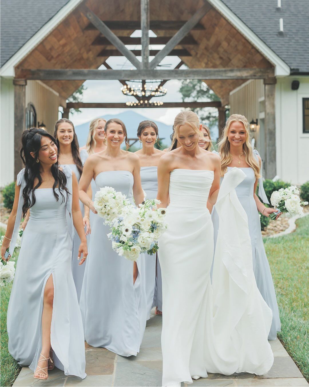 A bride in a white gown and bridesmaids in light blue dresses walk together on a path toward an outdoor venue entrance.