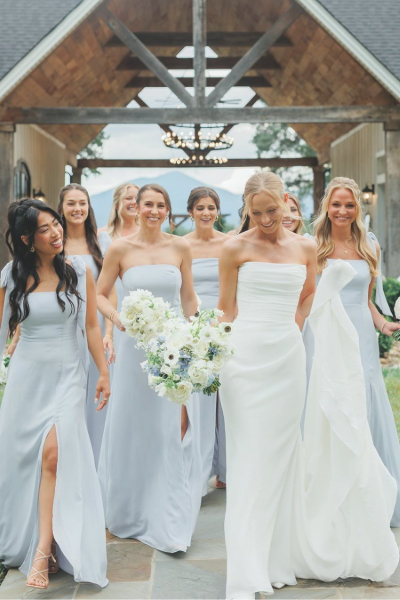 A bride in a white gown and bridesmaids in light blue dresses walk together on a path toward an outdoor venue entrance.