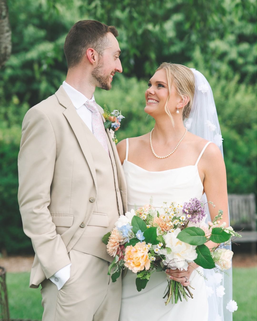 A smiling bride and groom in wedding attire look at each other while holding a colorful bouquet in an outdoor garden.