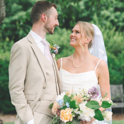A smiling bride and groom in wedding attire look at each other while holding a colorful bouquet in an outdoor garden.