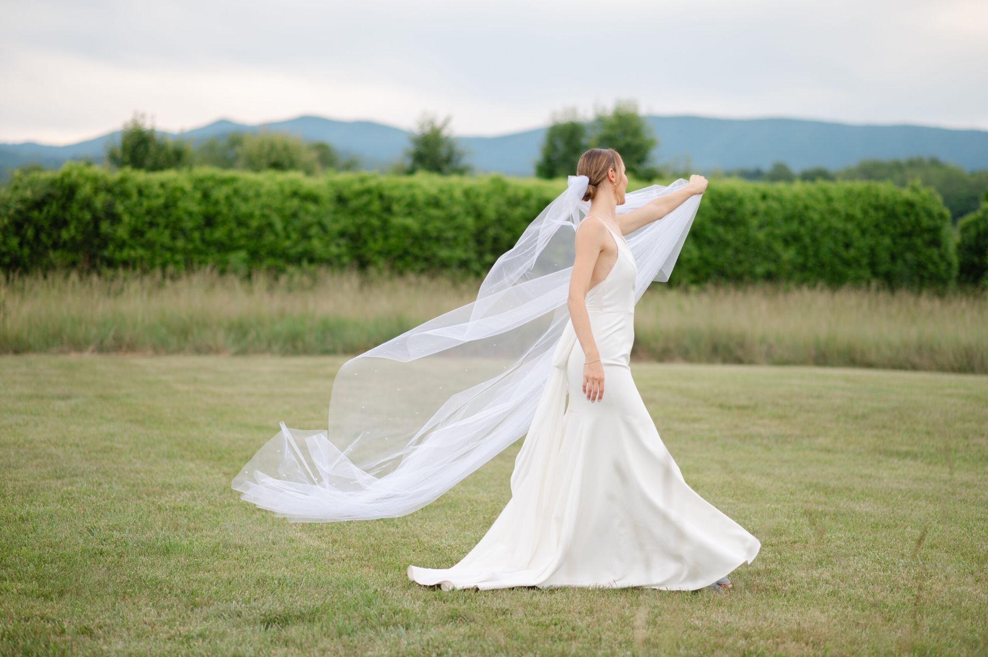 A person in a white wedding gown spins in a grassy field, their long veil billowing in the wind before distant hills.