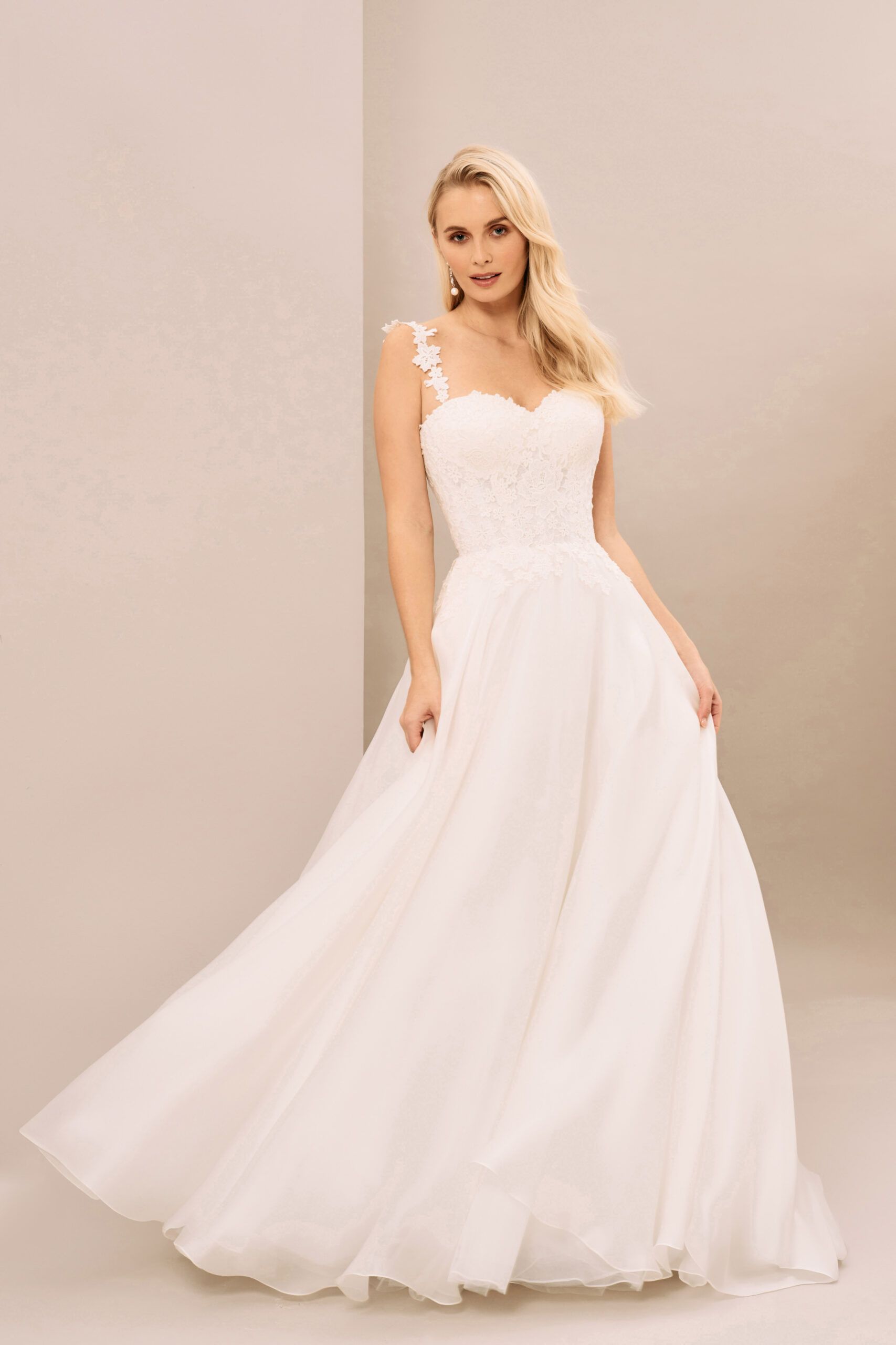 A person wearing a white, floor-length A-line wedding dress with a sweetheart neckline and floral strap details.