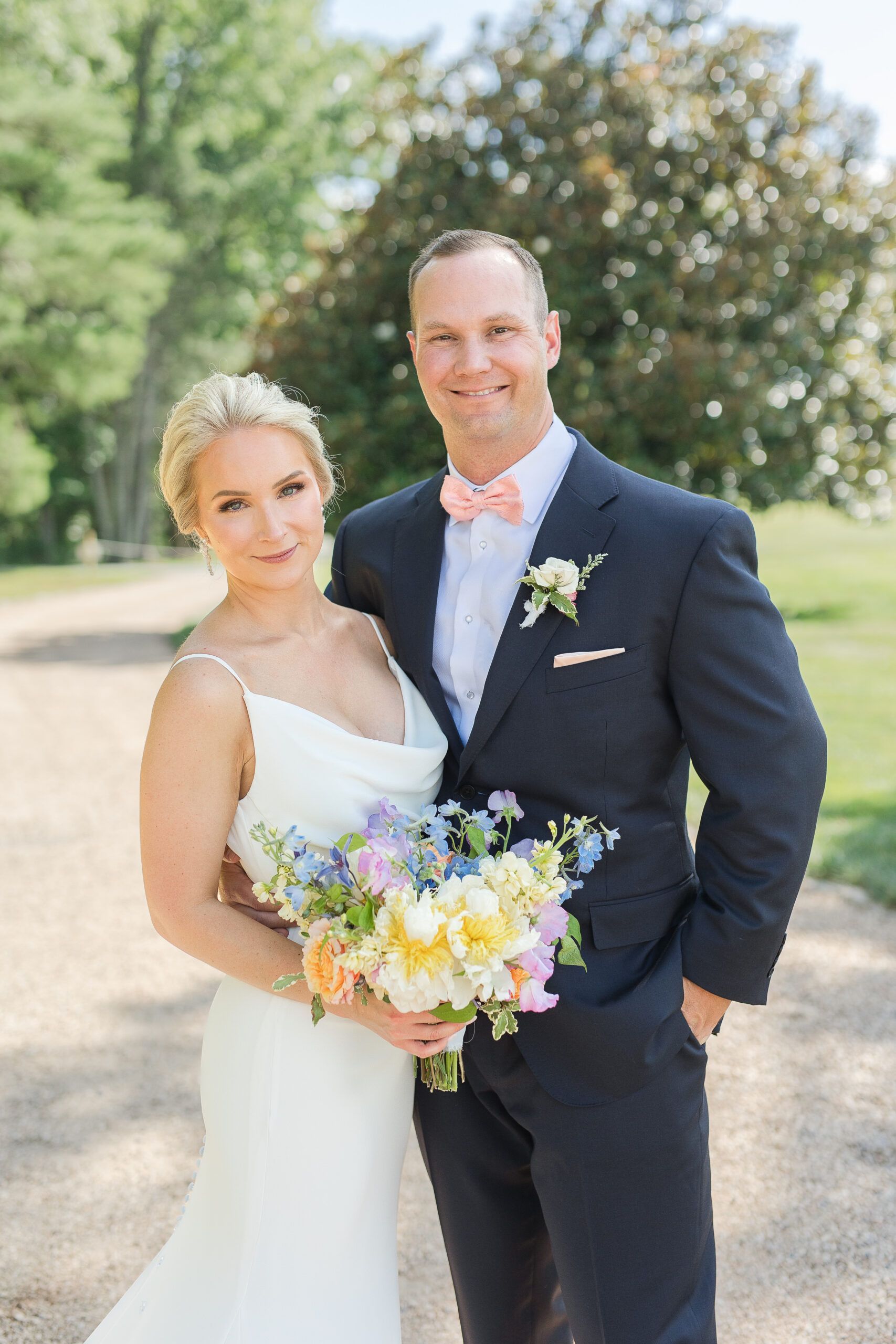 A bride in a white gown and a groom in a dark suit hold a floral bouquet together while standing on a gravel path.