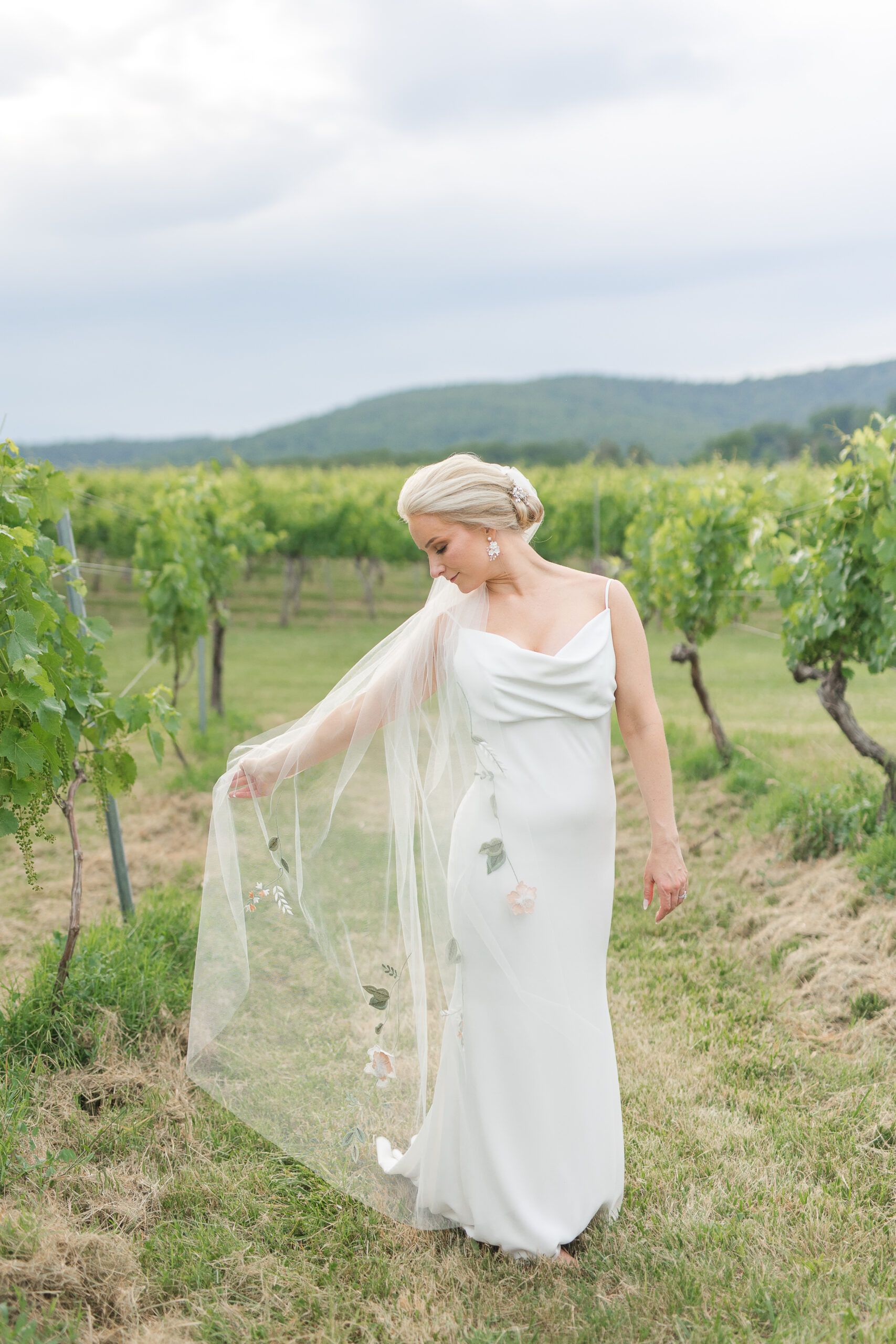 A person in a white wedding gown holding a delicate embroidered veil in a lush green vineyard under a cloudy sky.