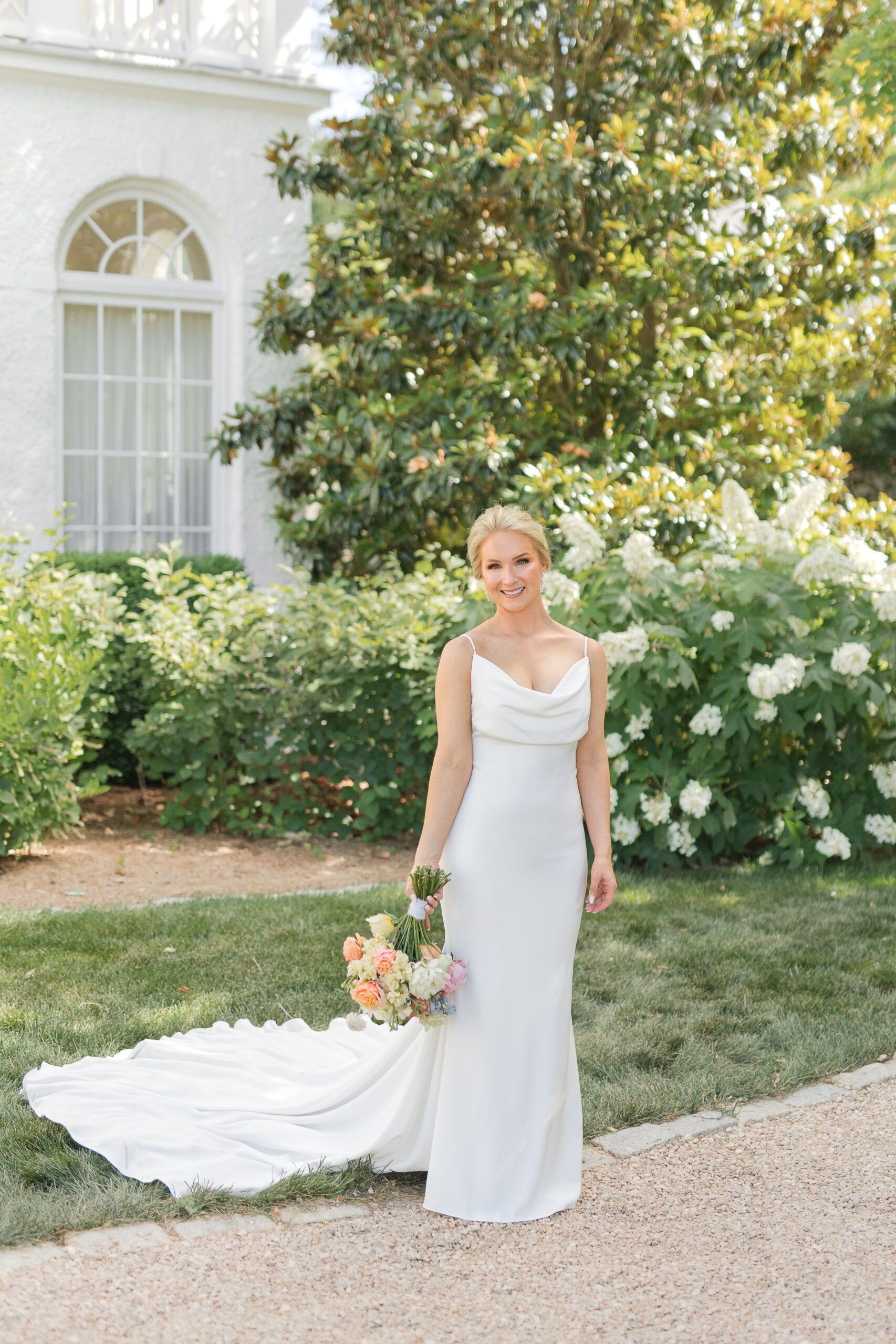 A person in a white wedding gown holding a floral bouquet, standing on a lawn in front of a white building and greenery.