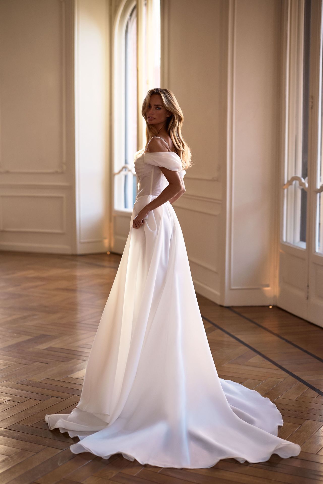 A person wearing an off-the-shoulder white A-line wedding dress with a train, standing in a brightly lit, elegant room.