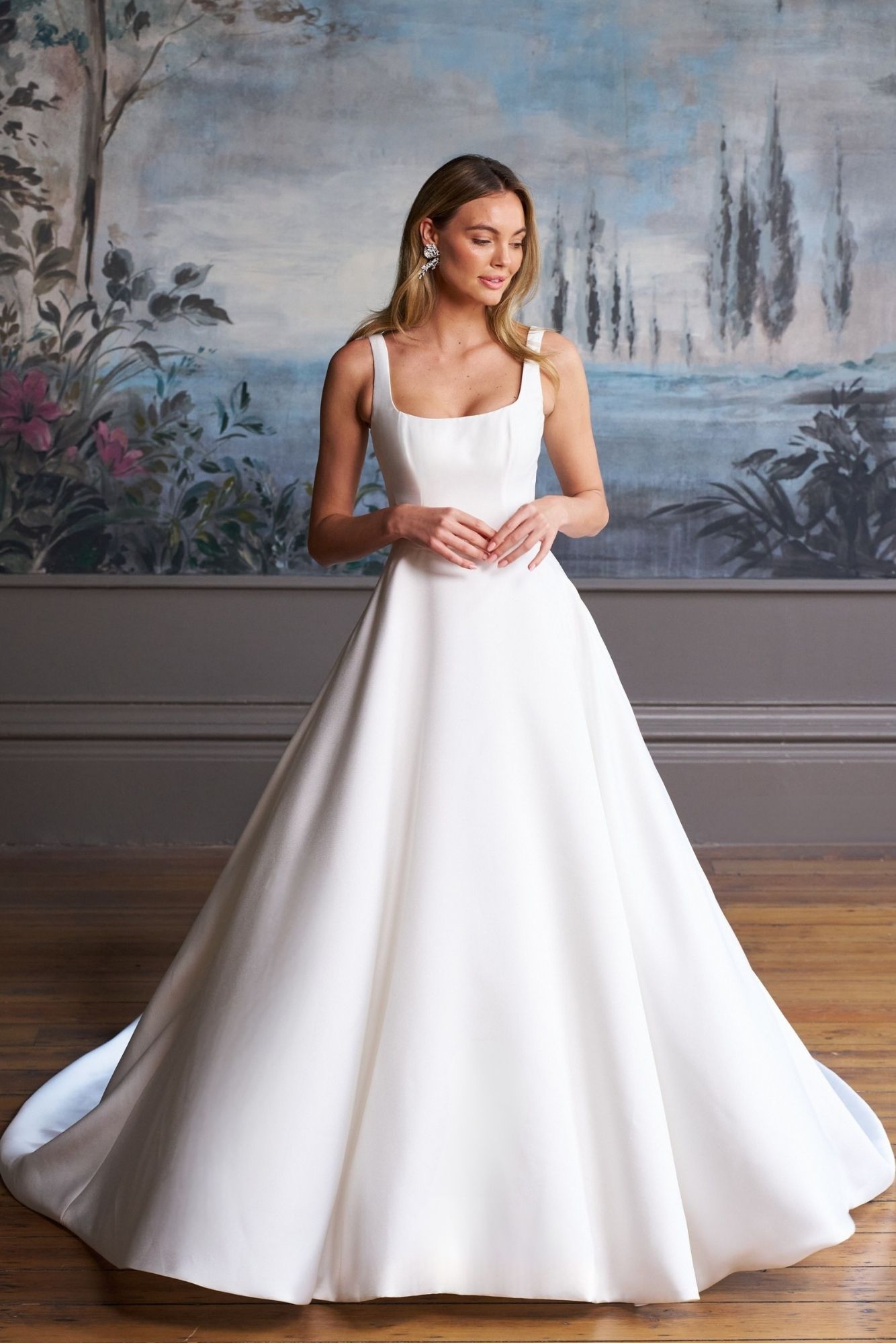 A model wearing a white, floor-length ballgown with a square neckline, standing in front of a painted wall mural.