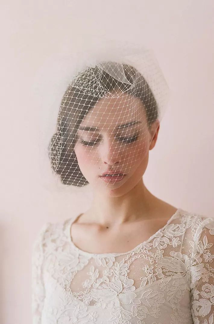 A person wears a vintage-style lace wedding dress and a birdcage veil with crystal accents over their face.