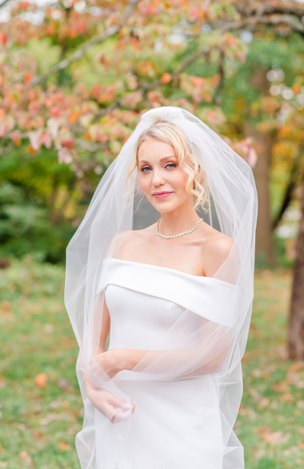 A bride in an off-the-shoulder white dress and veil poses in a park with autumn foliage in the background.
