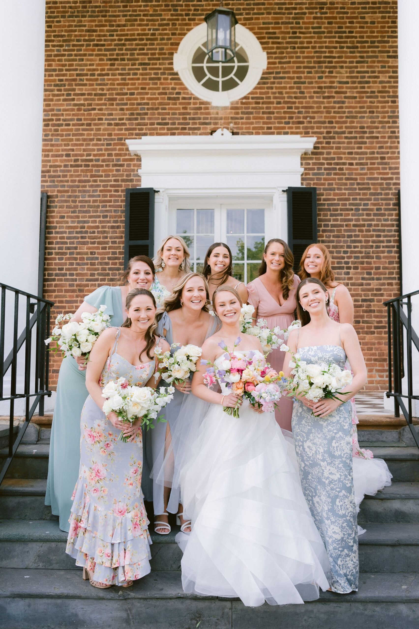 A bride in a white wedding gown and seven bridesmaids in varied floral and solid-colored dresses pose on brick steps.