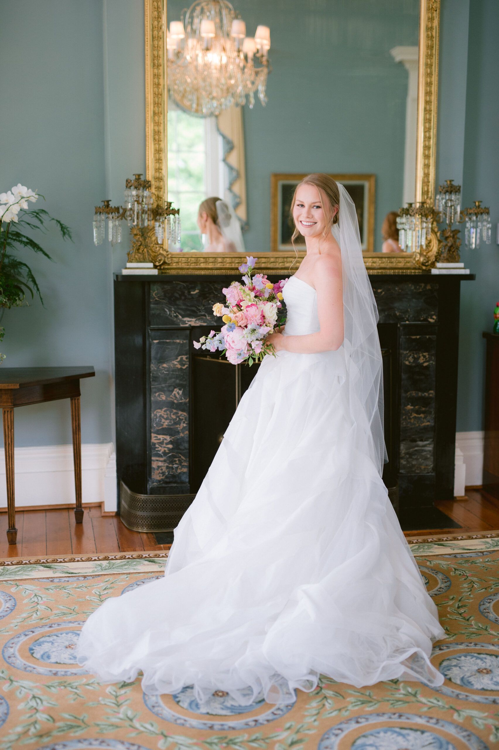 A smiling bride in a white wedding gown and veil, holding a bouquet in a room with a fireplace and large mirror.