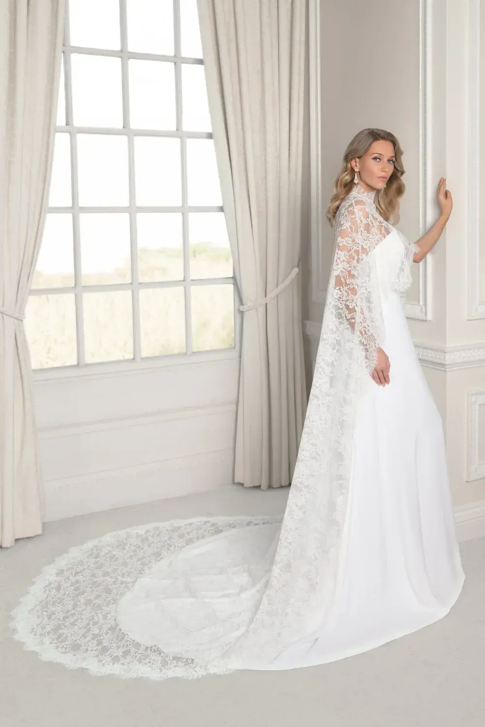 A person in a white wedding gown with long lace sleeves and a matching lace cape, standing near a window with curtains.