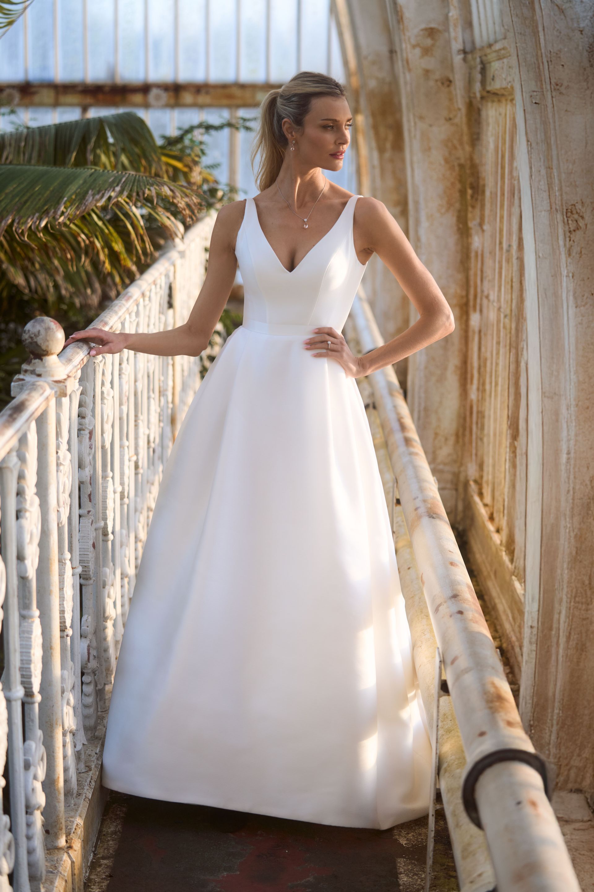 A person wears a minimalist white wedding dress with a V-neckline, posing on a sunlit conservatory balcony.
