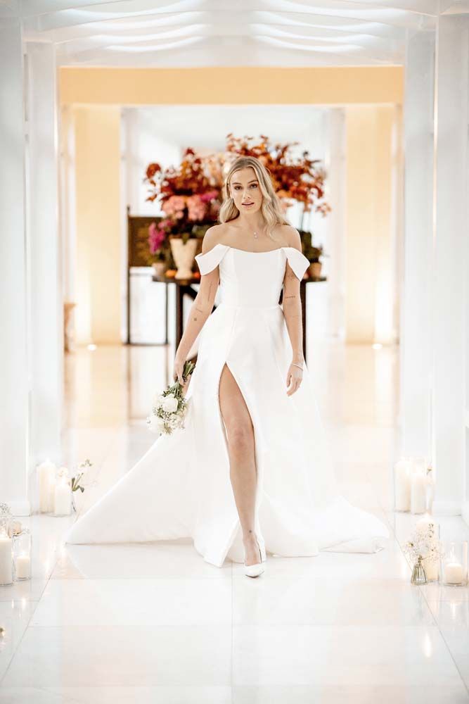 A person in a white off-the-shoulder wedding gown with a high slit walks down a bright, columned indoor hallway.