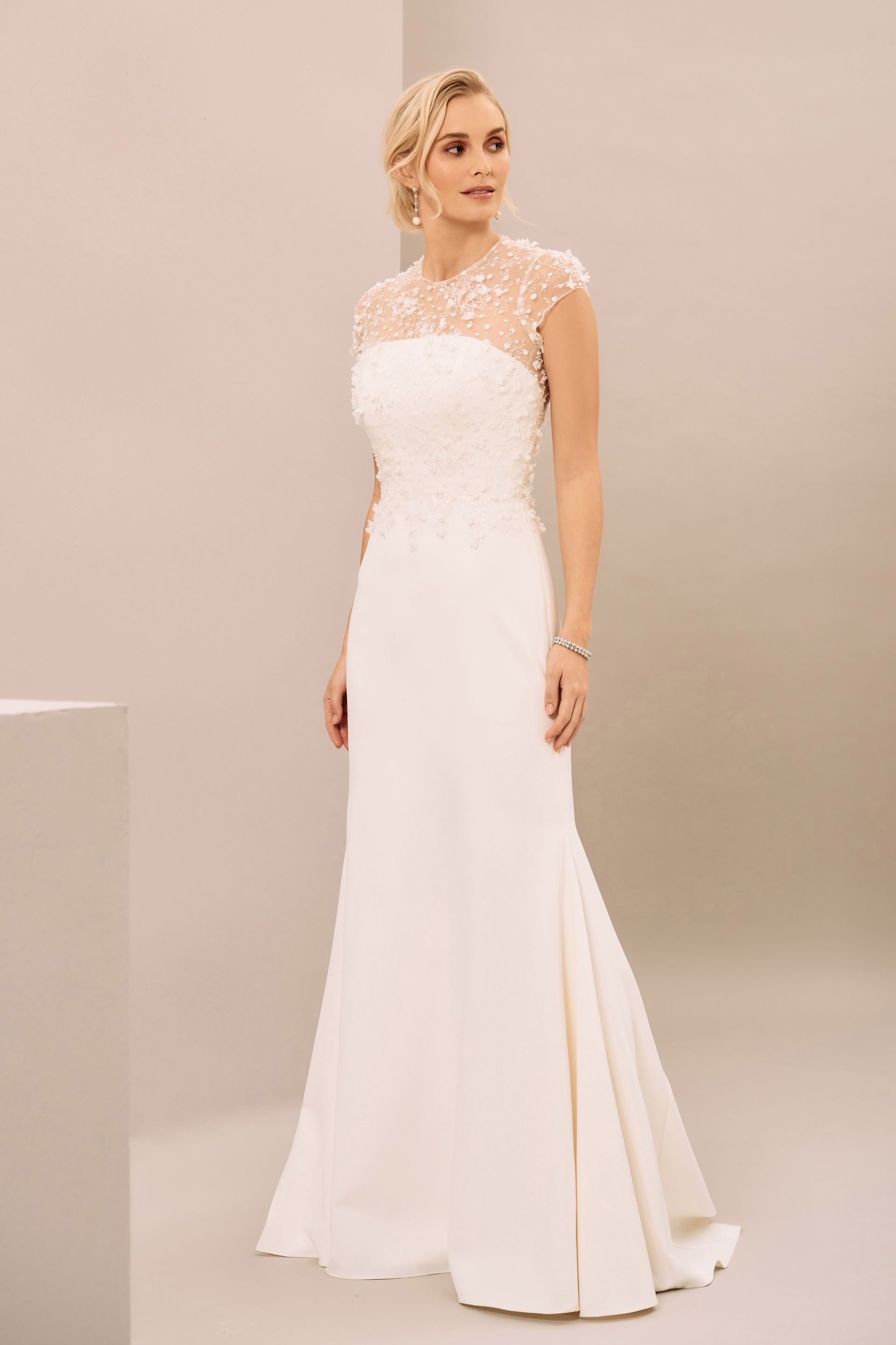 A woman wearing a floor-length white wedding dress with a lace-detailed bodice and cap sleeves against a neutral background.