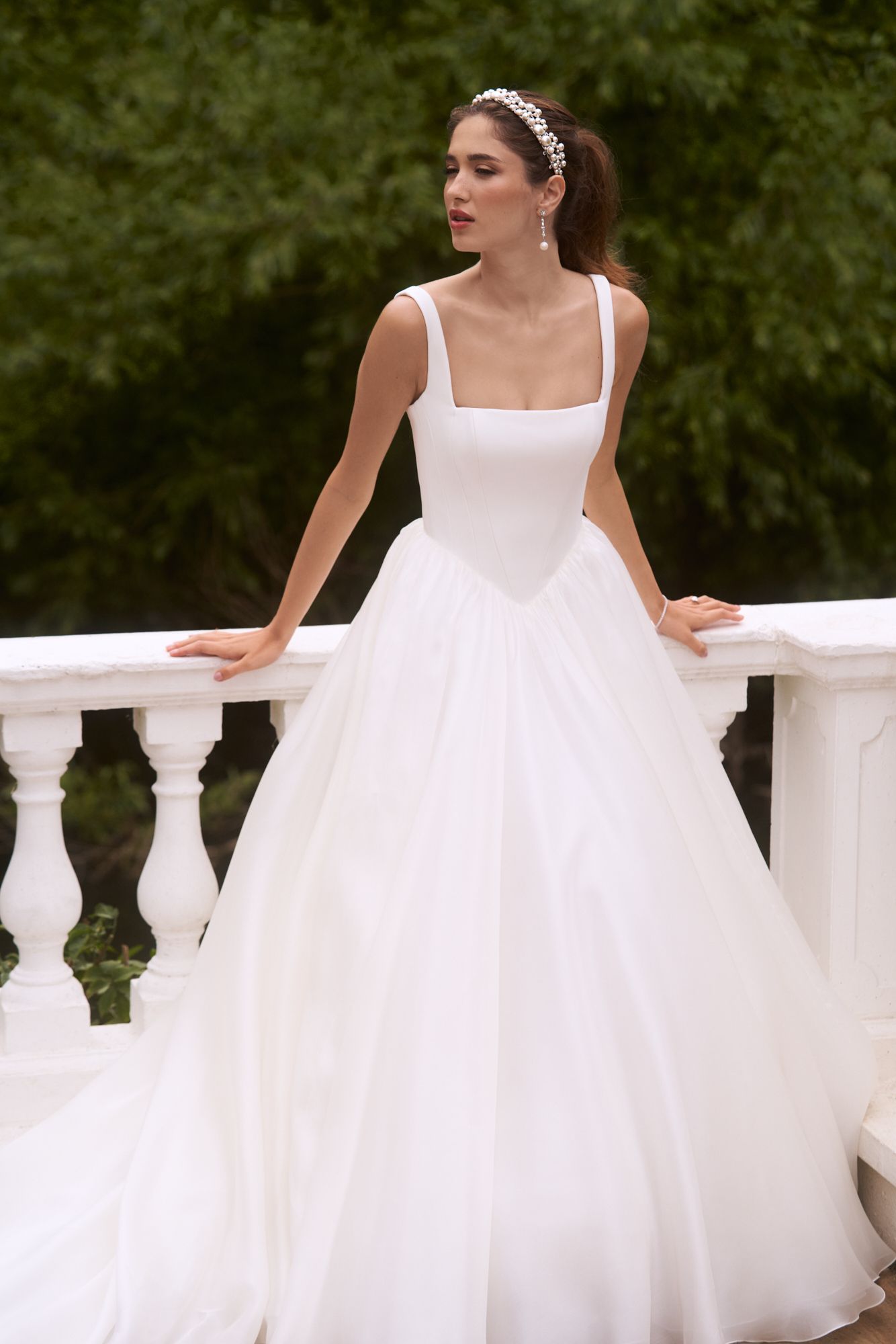 A person in a white wedding gown with a square neckline and full skirt stands against a white railing in a garden.