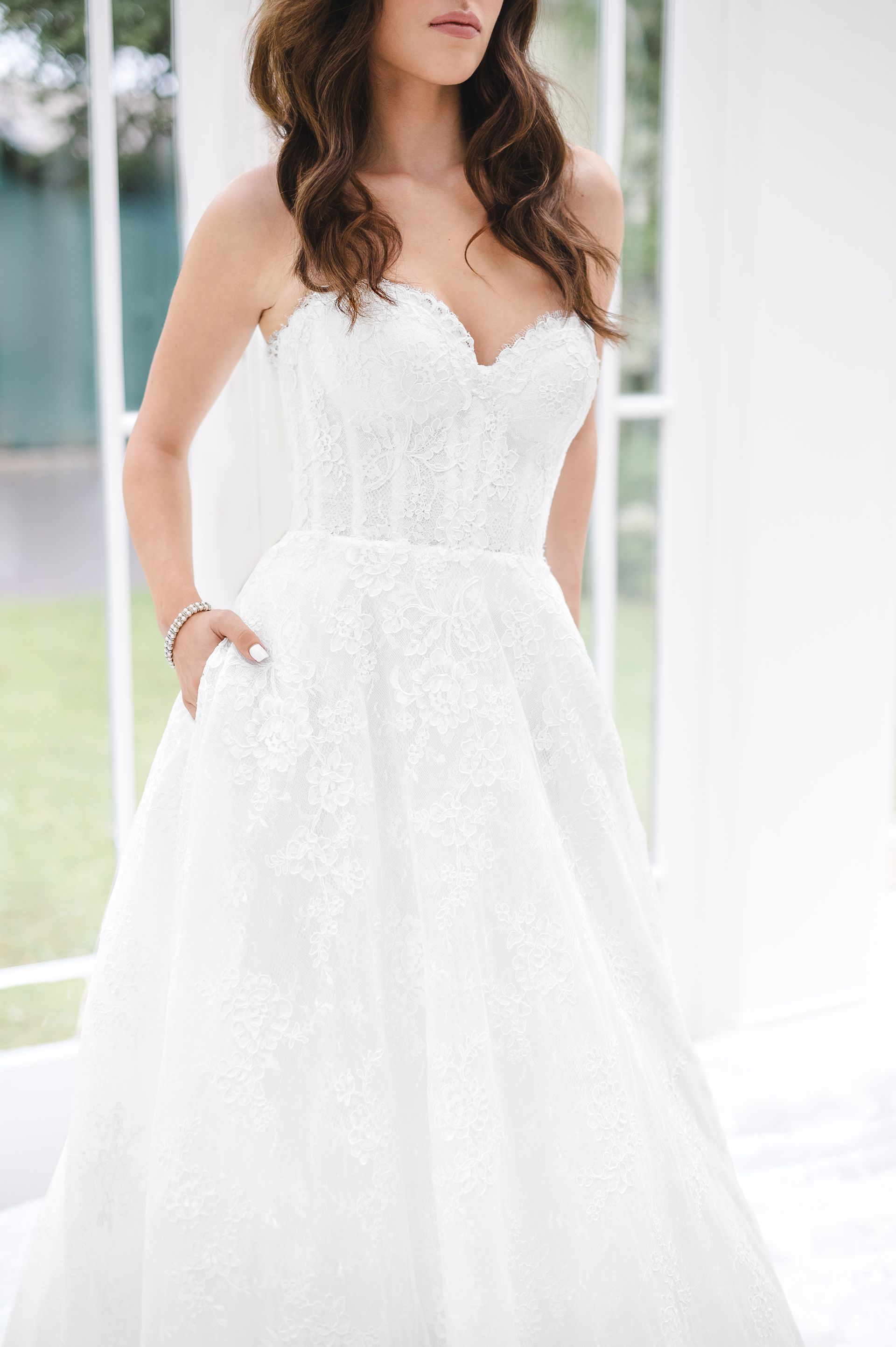 A person wearing a white, strapless, lace-textured wedding gown with pockets, standing indoors.