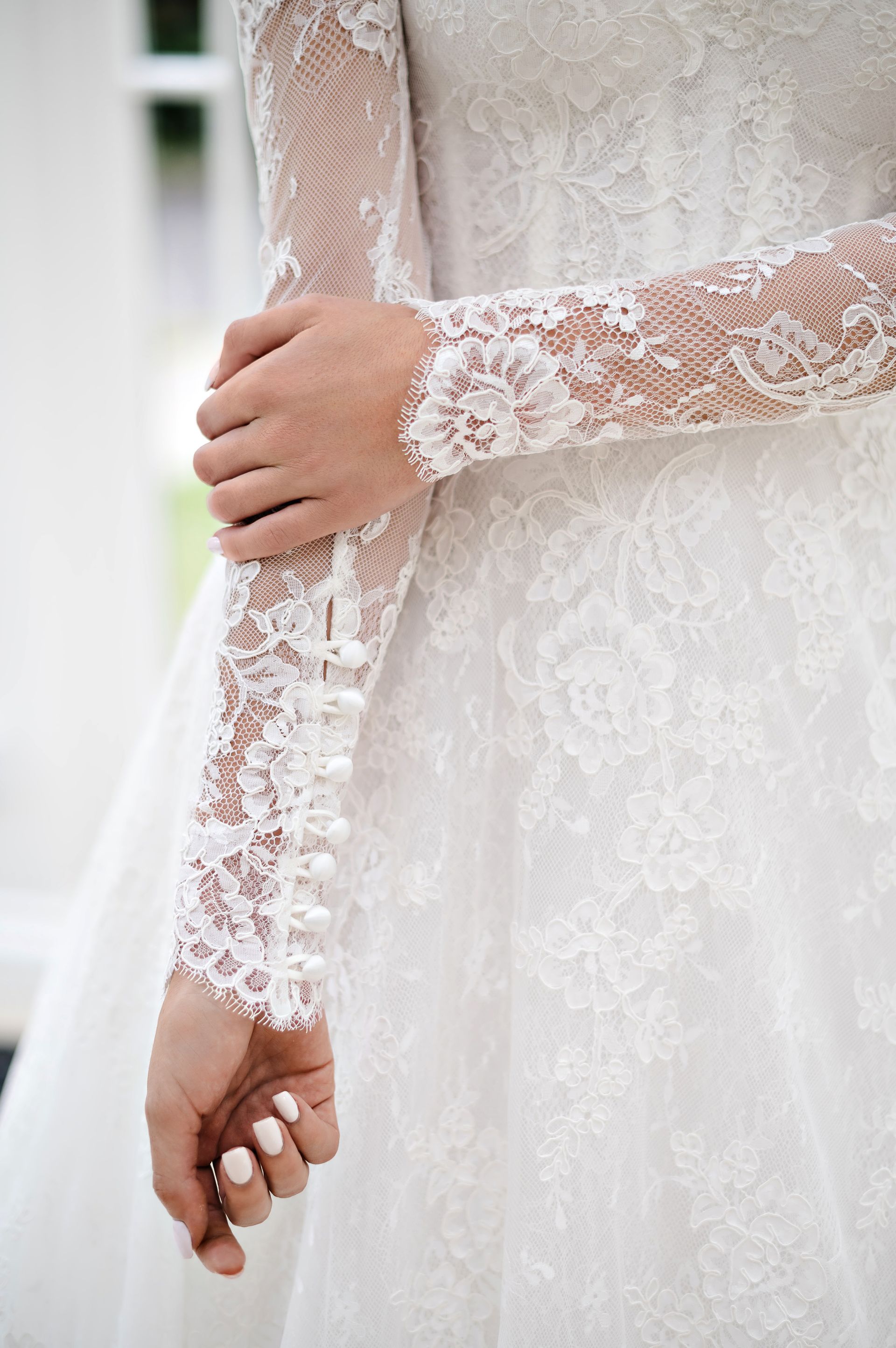 Close-up of a person's arms and hands wearing a long-sleeved white lace bridal gown with button details at the wrists.