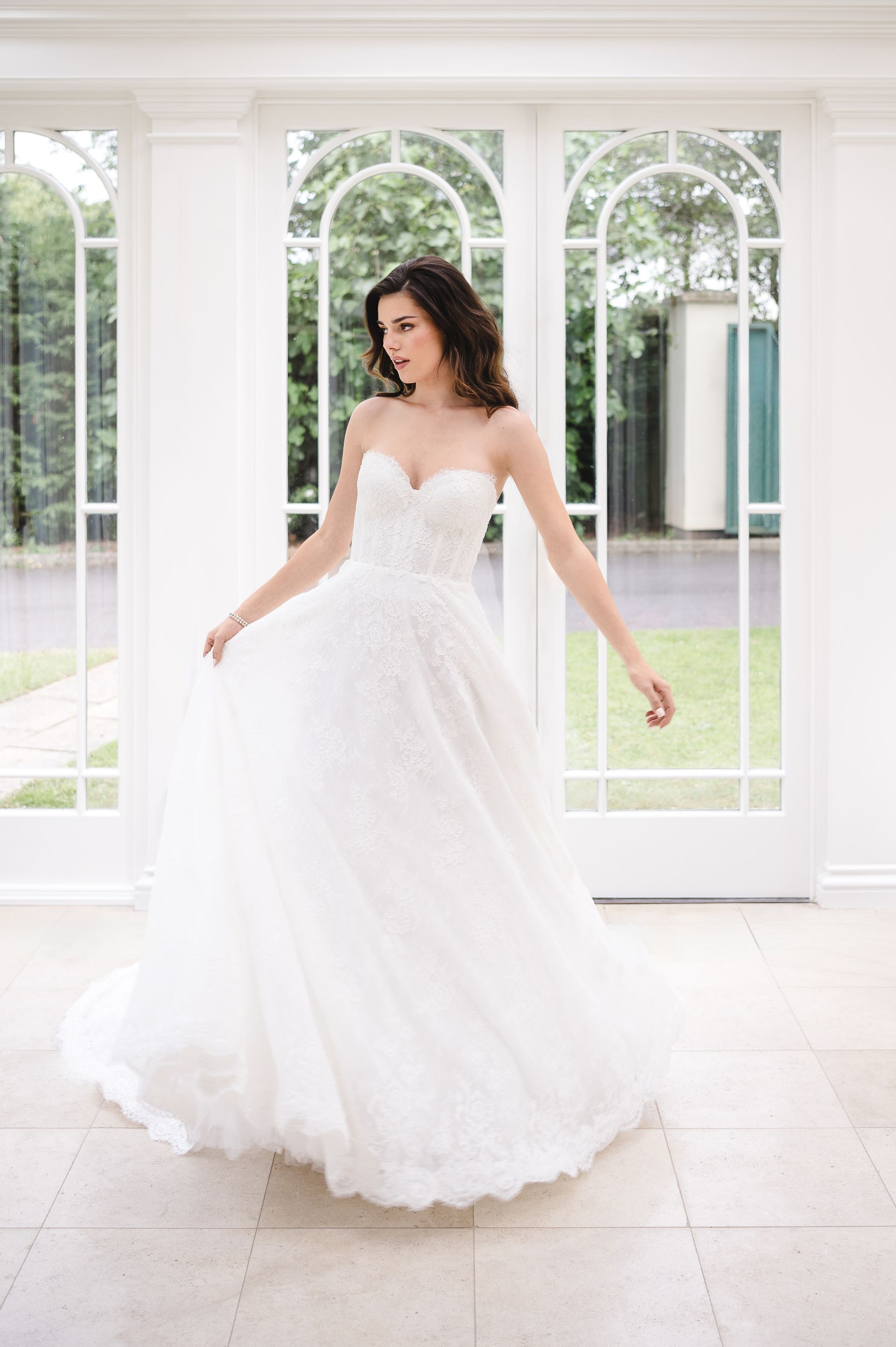 A person in a white strapless wedding gown with a full, textured skirt twirls inside a bright room with glass doors.
