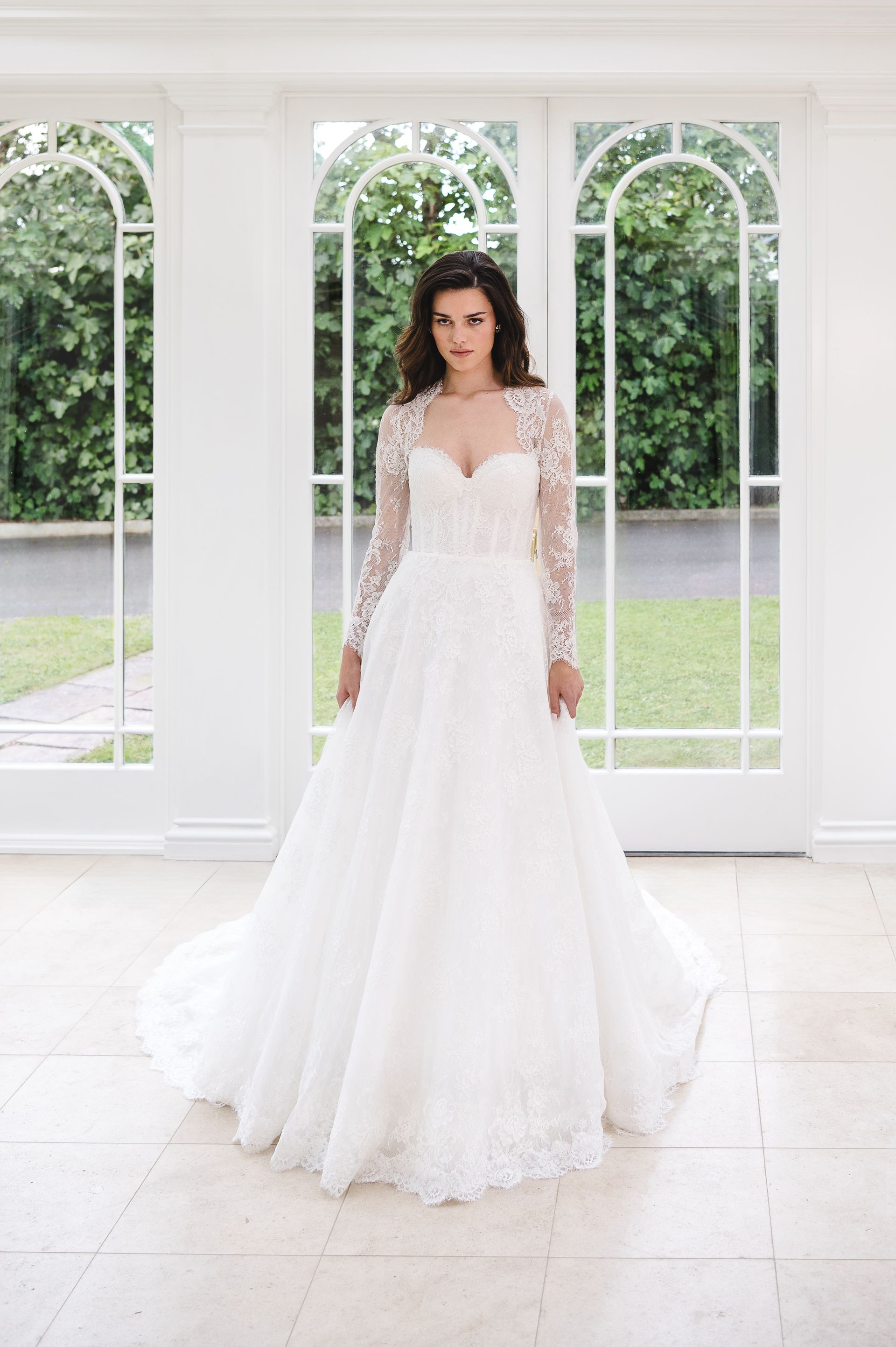 A person wears a white, floor-length lace wedding dress with long sleeves, standing in a bright sunroom with arched glass.