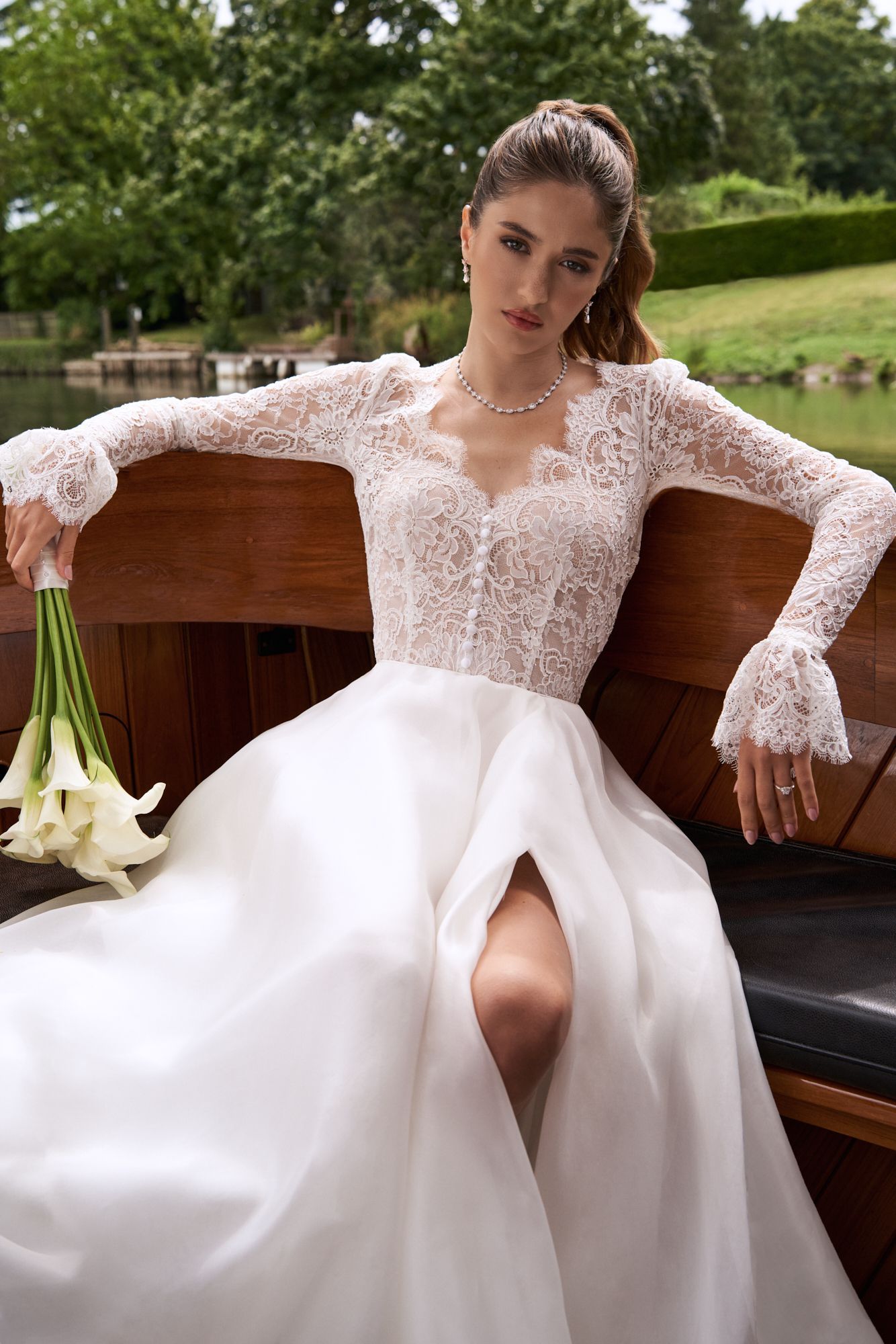 A person in a lace-sleeved wedding dress with a leg slit holds calla lilies while sitting on a wooden boat outdoors.