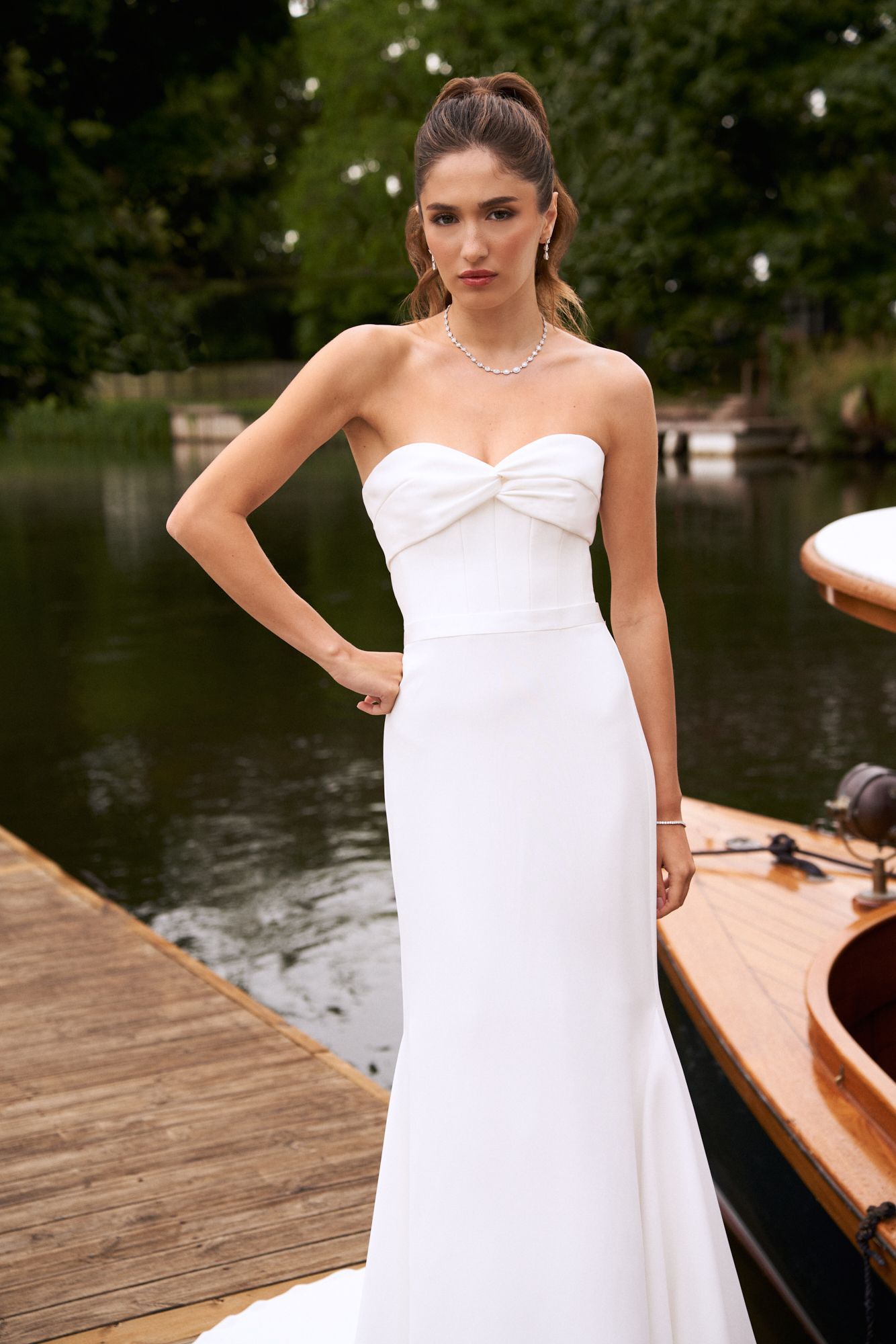 A person wearing a strapless white wedding gown with a sweetheart neckline stands on a dock next to a boat.