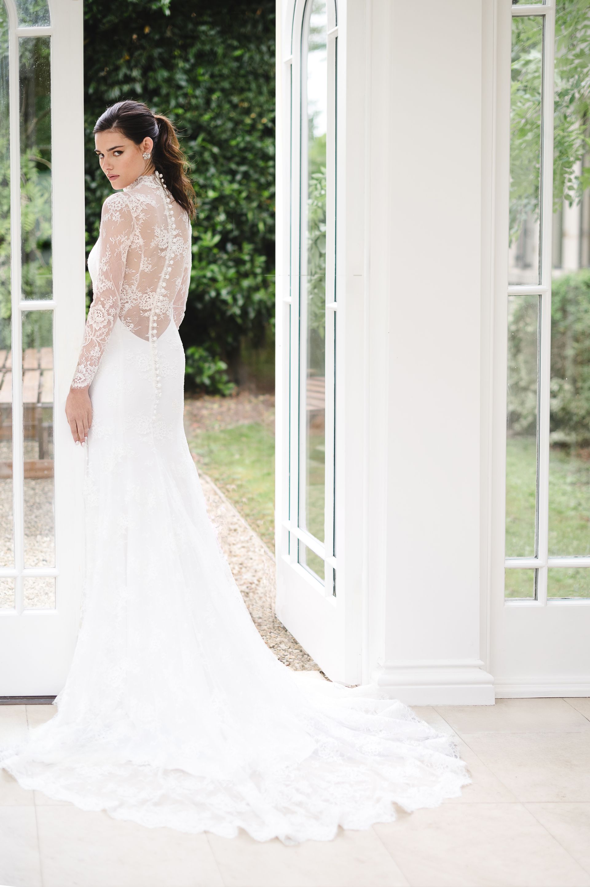 A person wearing a long-sleeved, lace-back white wedding dress standing in an open doorway.