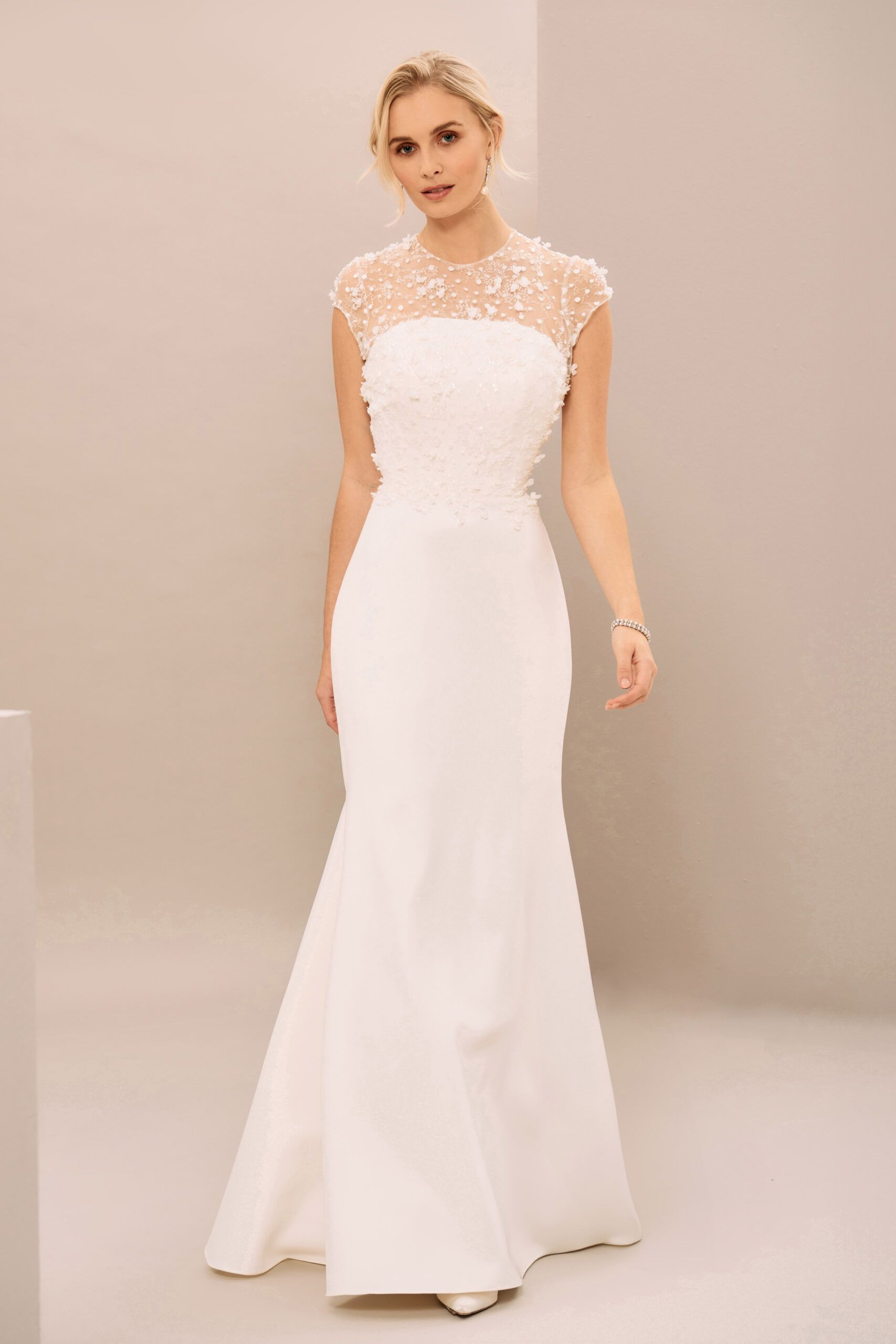 A model wears a form-fitting white floor-length bridal gown with a high-neck lace illusion bodice against a plain background.