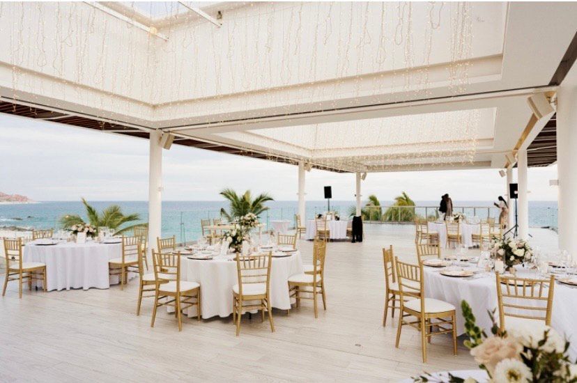 Wedding reception setup on a white terrace overlooking the ocean, featuring round tables with gold chairs and flowers.