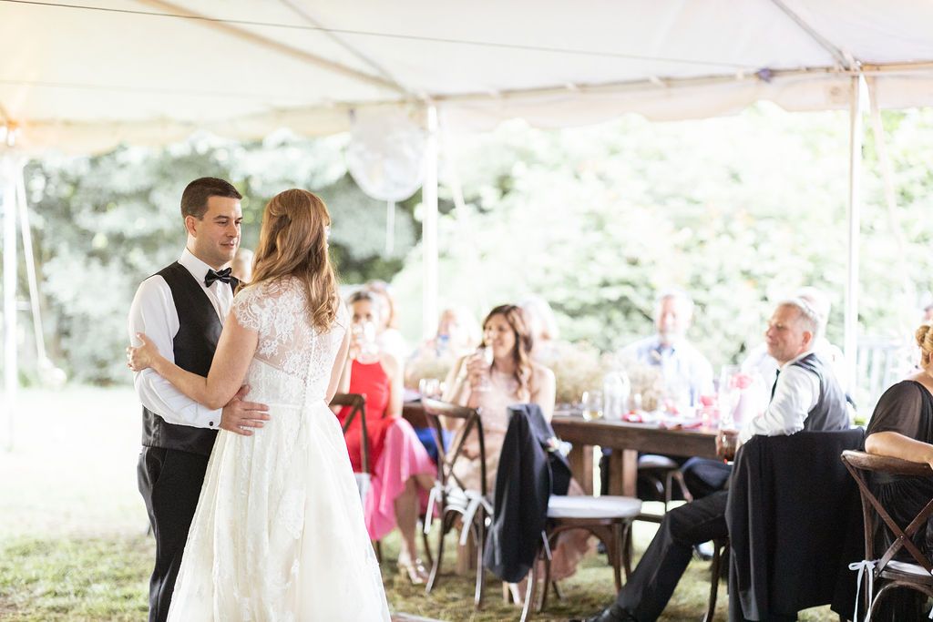 A bride and groom dancing at an outdoor reception under a white canopy tent, with guests seated at tables in the background.