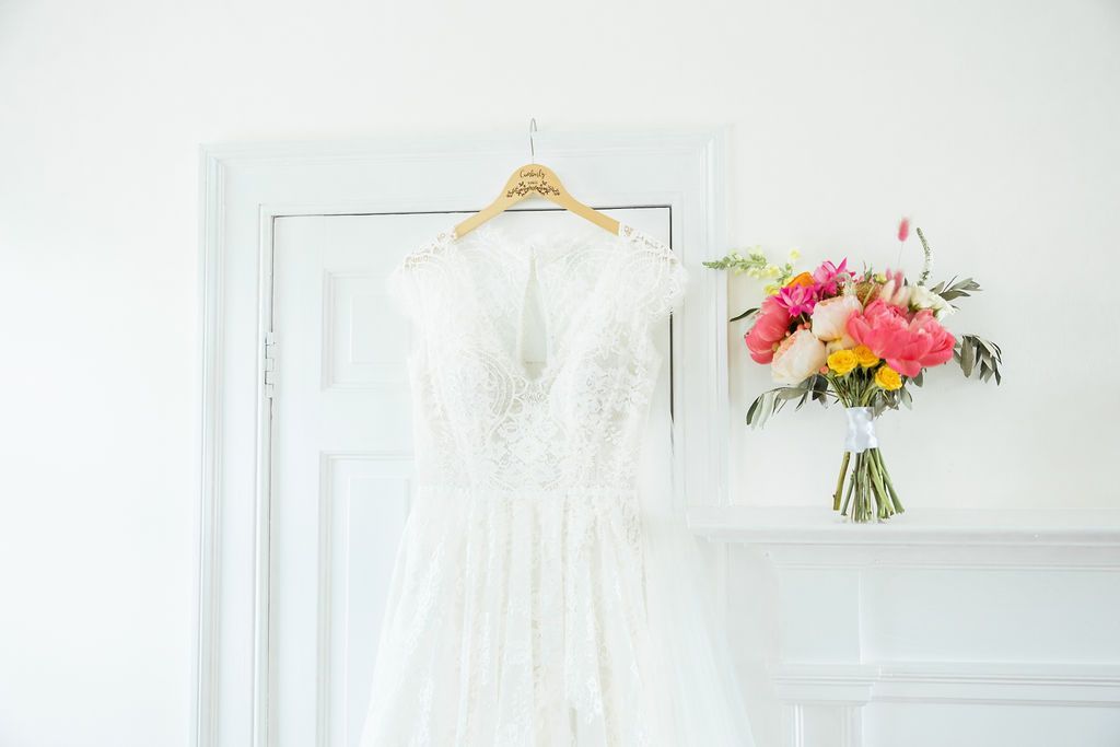 A white lace wedding dress hangs on a hanger in front of a white door next to a vibrant bouquet of pink and yellow flowers.