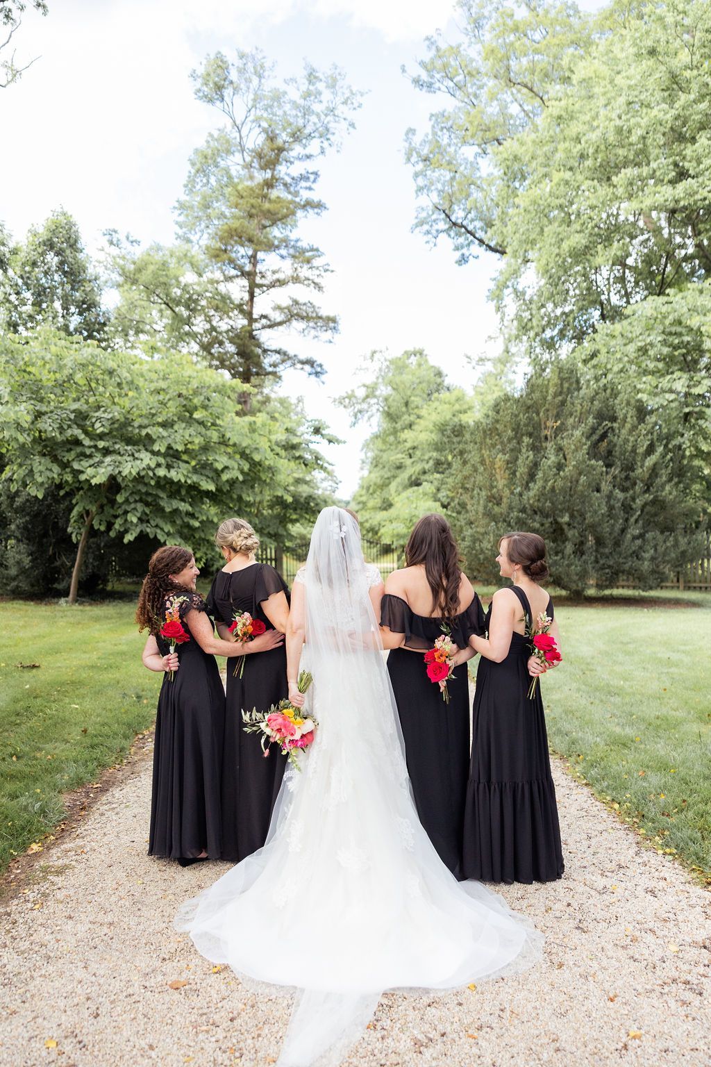 A bride in a white gown and four bridesmaids in black dresses standing on a gravel path outdoors holding colorful bouquets.