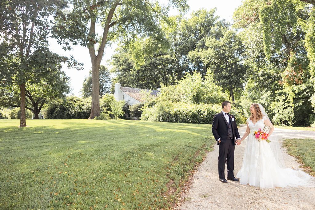 A wedding couple stands together on a gravel path in a lush green park with a large house visible in the background.