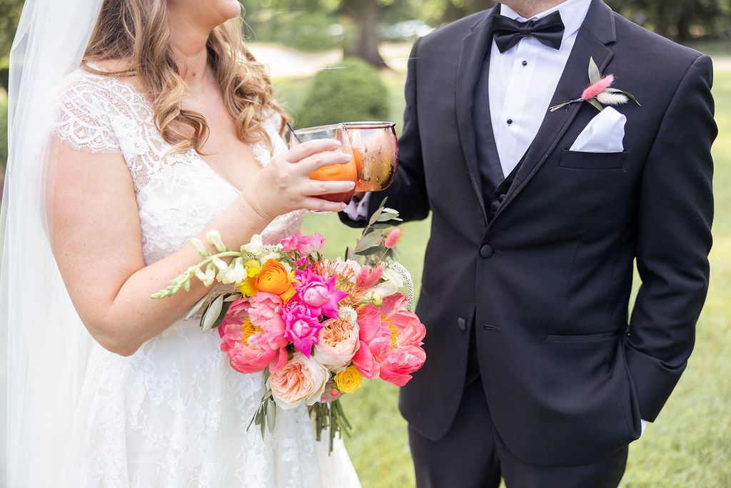 A bride and groom in formal wedding attire holding cocktails and a vibrant bouquet of pink, orange, and yellow flowers.